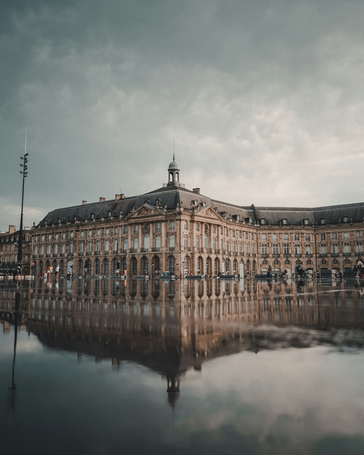 La place de la Bourse et son dôme se reflètent dans la fine nappe d’eau du Miroir d’Eau, sous un ciel nuageux