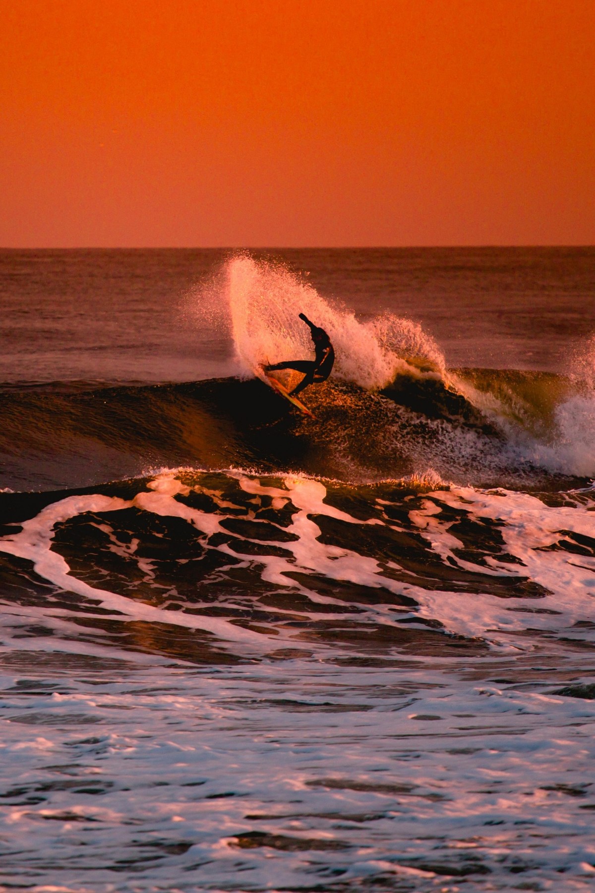 Surfeur en pleine figure au coucher du soleil : gerbe d'eau rosie par le ciel orangé, océan et écume au premier plan