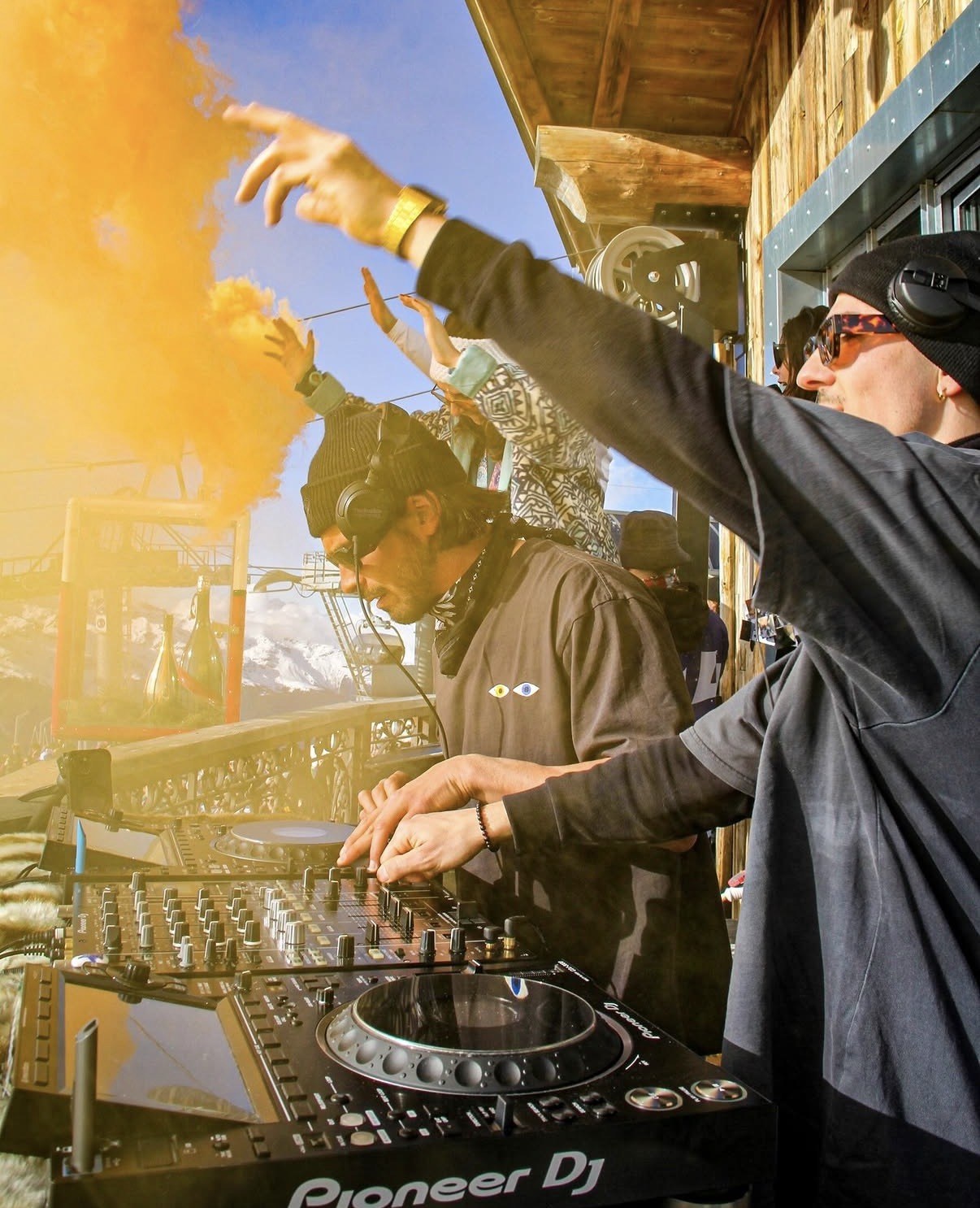 Deux DJs mixent sur une terrasse en bois face aux sommets enneigés, un fumigène orange sur la gauche