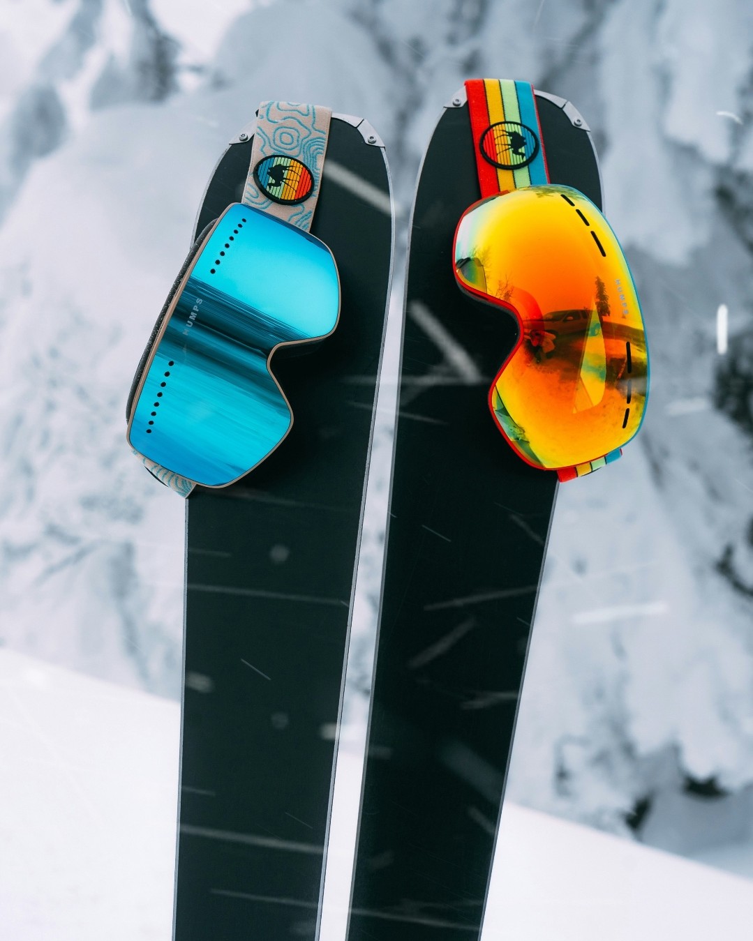 Deux skis noirs plantés dans la neige, chacun coiffé d'un masque de ski, l'un à verre bleu turquoise, l'autre à verre orangé, sur fond de sapins enneigés